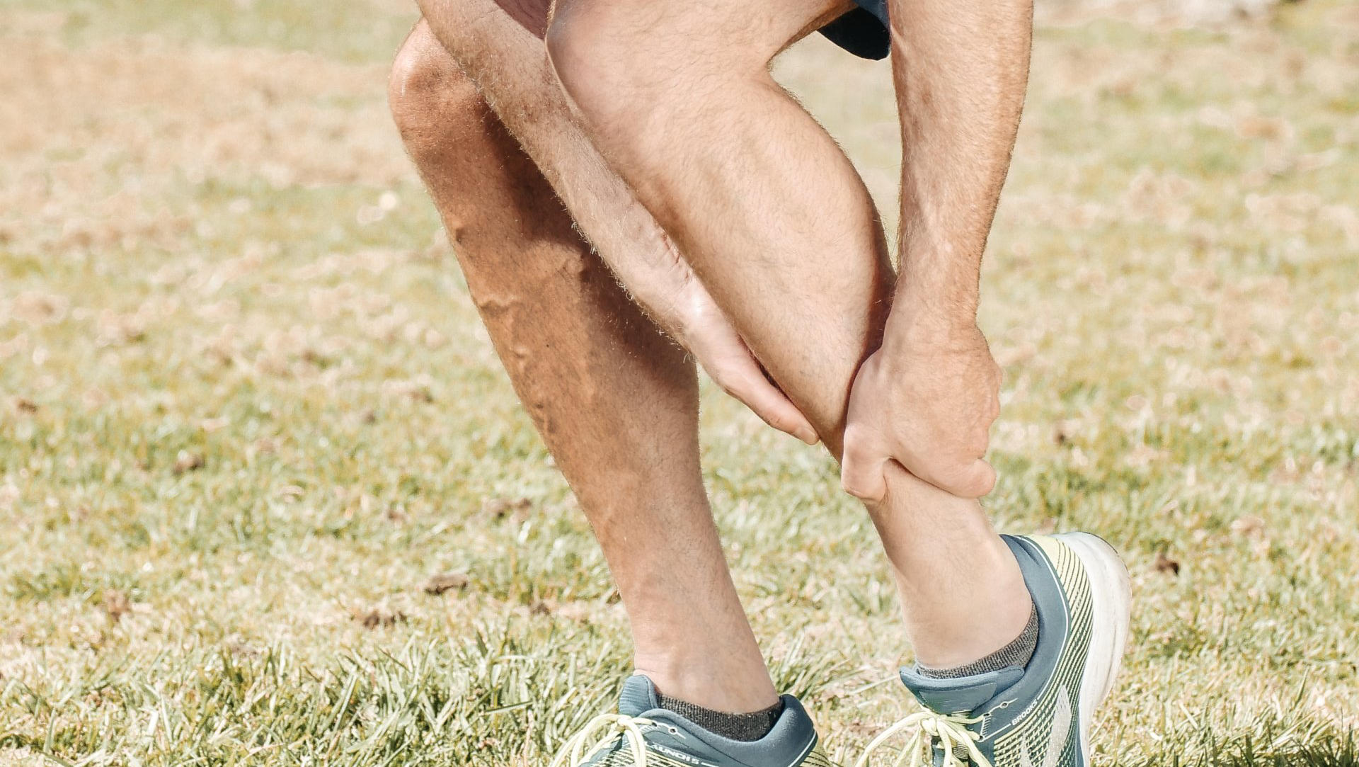 Man holding his lower leg, possibly in pain from plantar fasciitis. Green grass, blue sneakers.