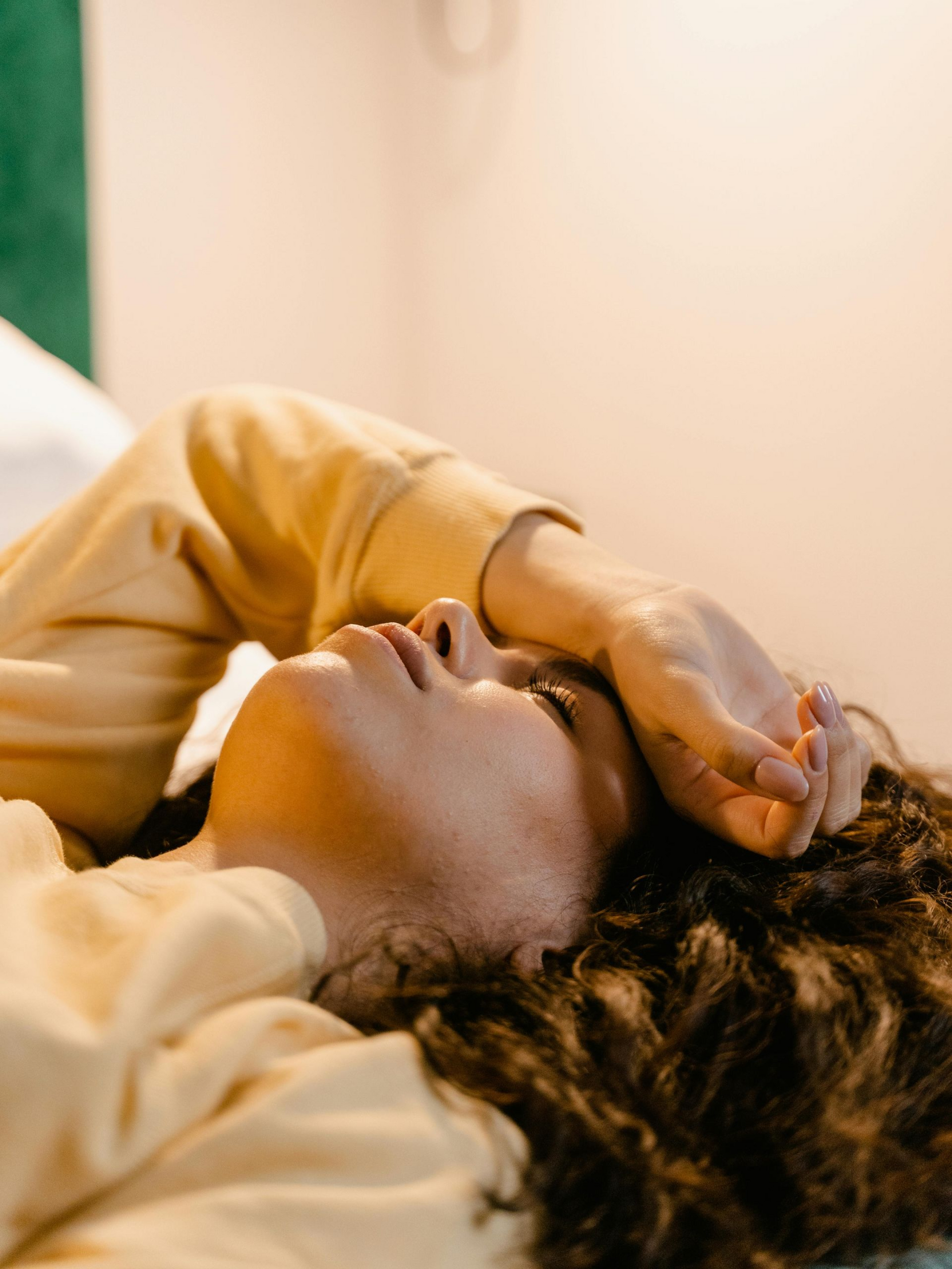 A person with curly hair wearing a yellow sweatshirt rests with their hand over their forehead while lying down.