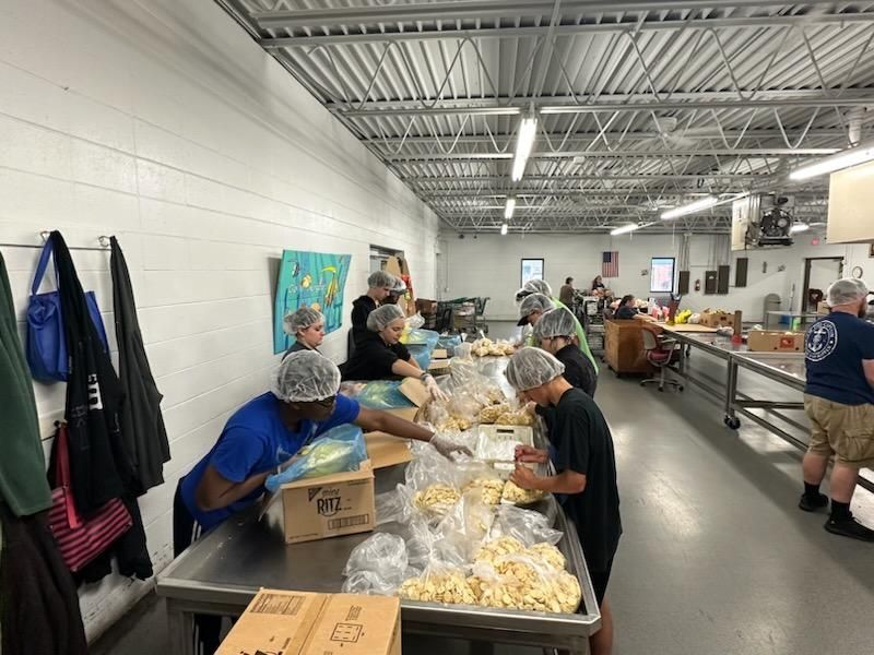 People in hairnets packaging food at a facility with stainless steel tables, under fluorescent lights.