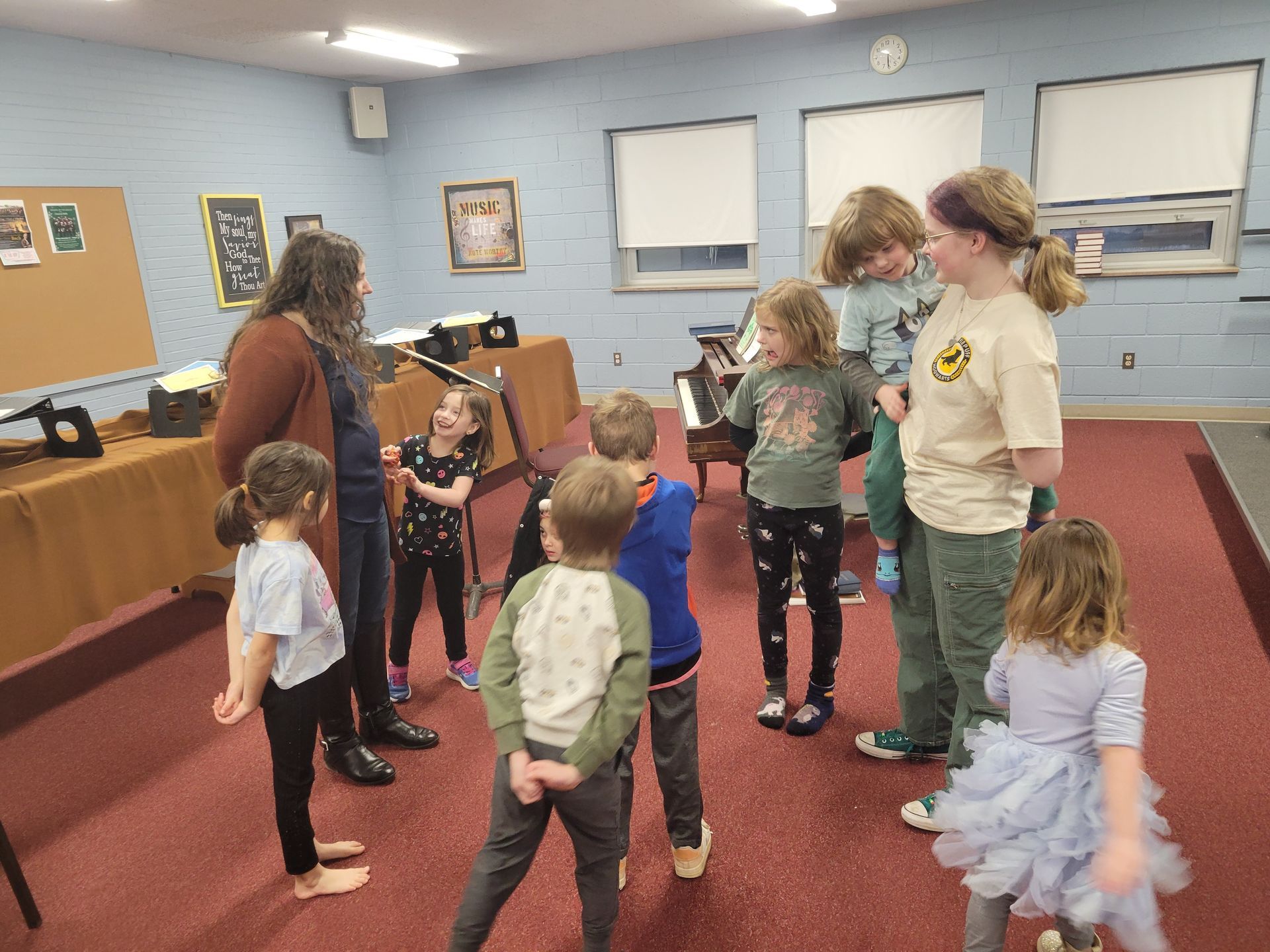 Children and an adult in a music room, possibly a lesson. They are gathered in a circle near a piano.
