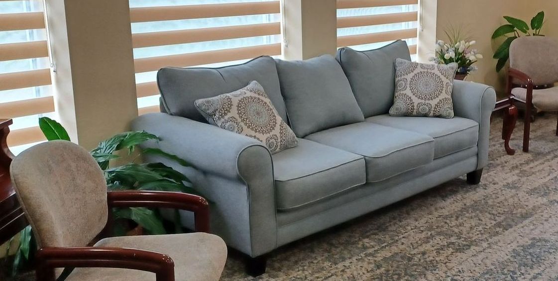 Blue sofa with two patterned pillows, a chair, and a plant next to a window with blinds.