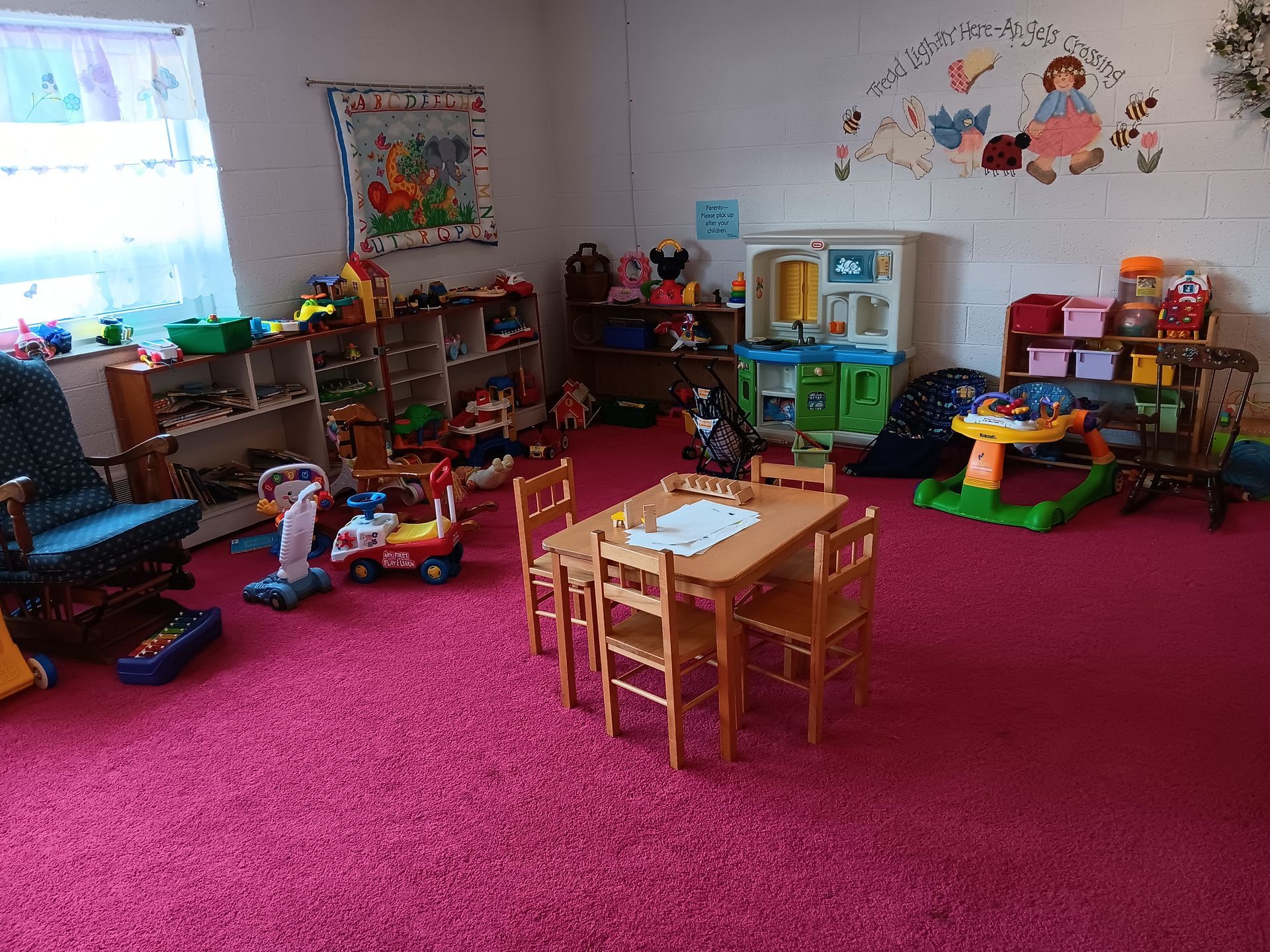 A playroom with pink carpet, toys on shelves, small table and chairs, and a window.