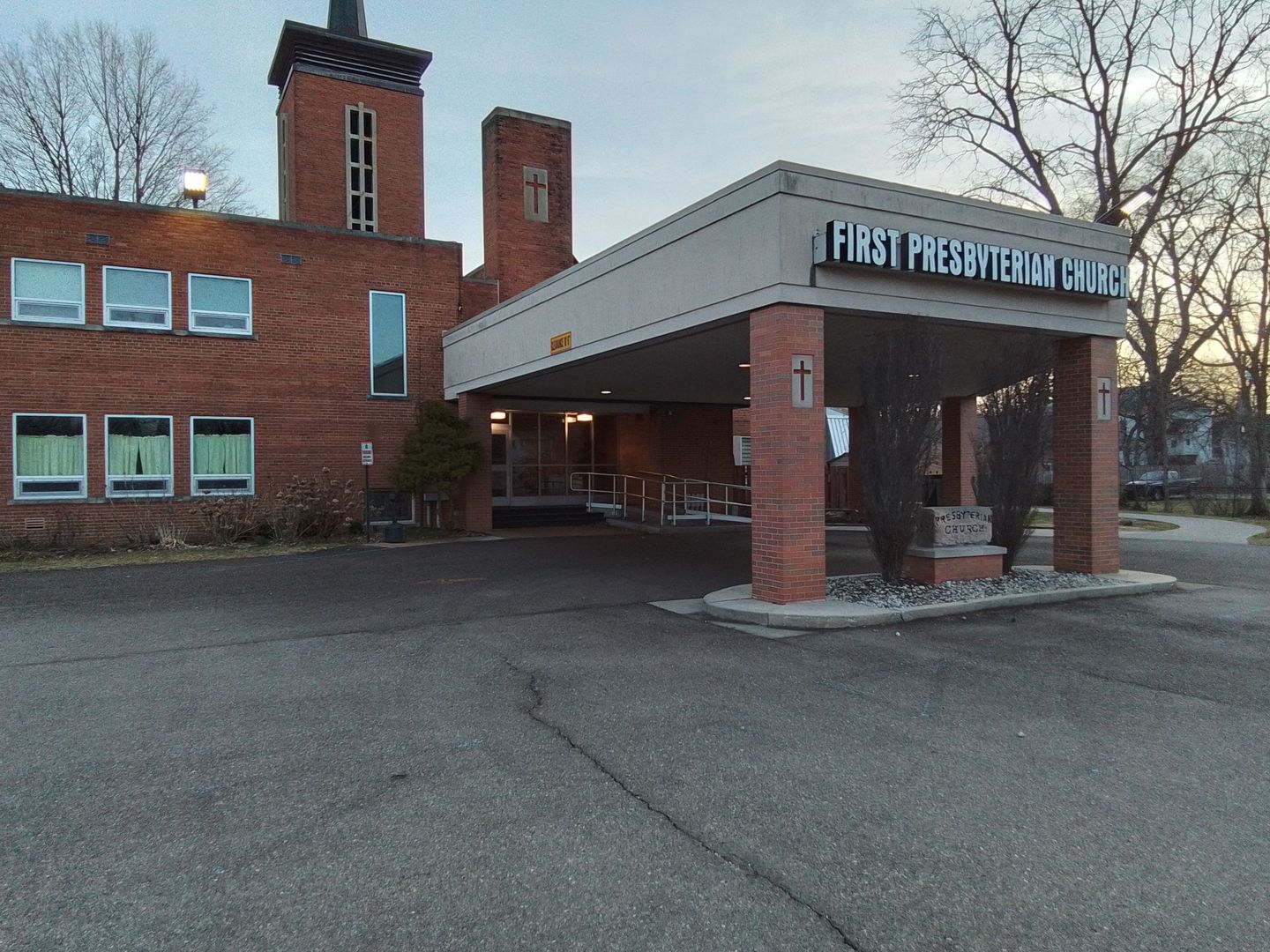 Exterior view of First Presbyterian Church building with a covered entrance. Red brick, gray awning, and parking area.
