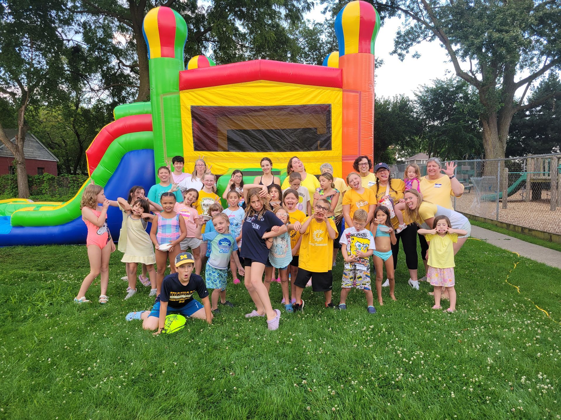 Group of children and adults near a colorful inflatable bounce house and slide on a grassy lawn.