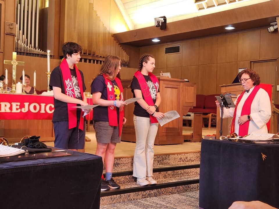 Three people stand with a minister at a church altar. They wear red stoles and hold papers, presumably for a ceremony.
