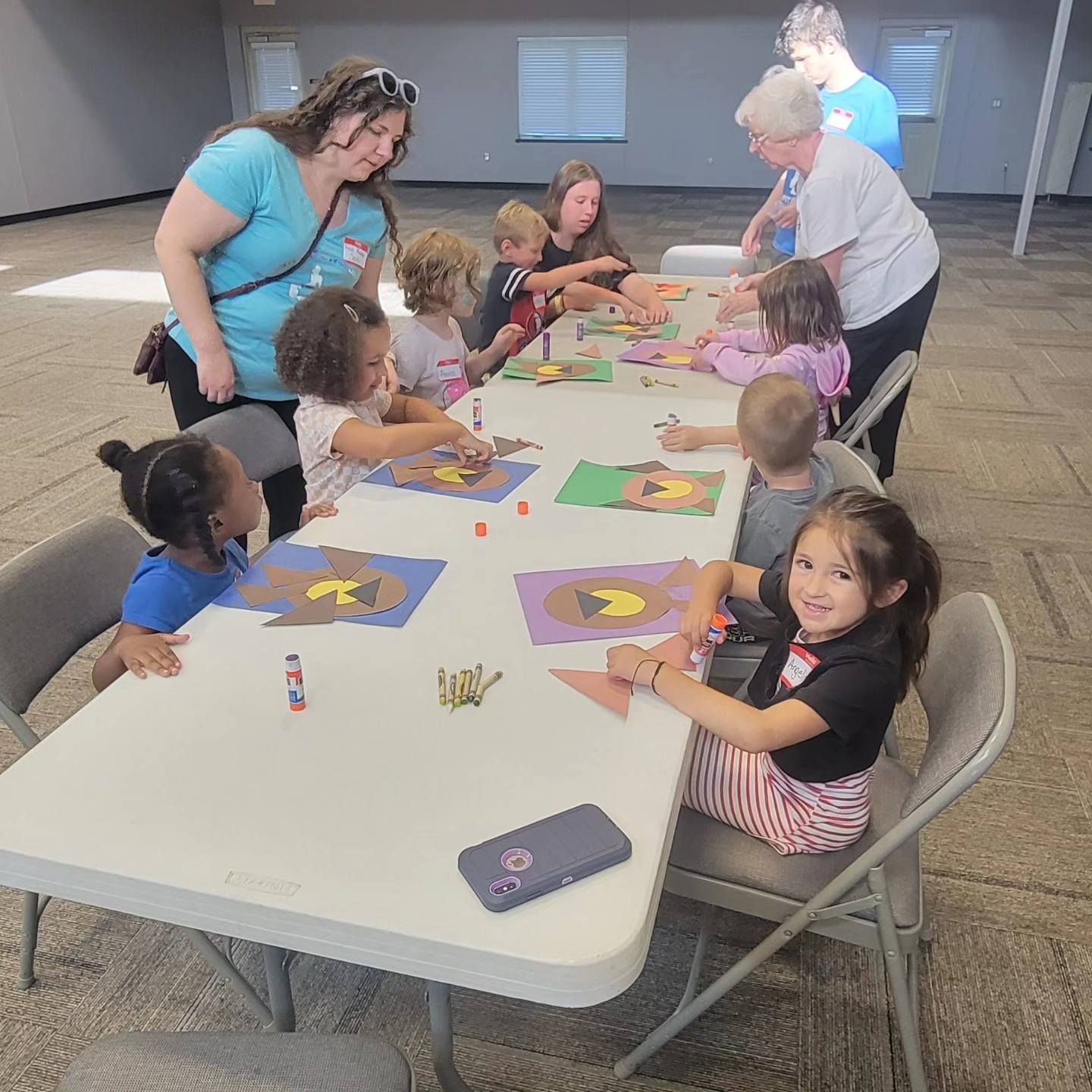 Children at a table doing crafts, adults assisting, indoors, daylight.