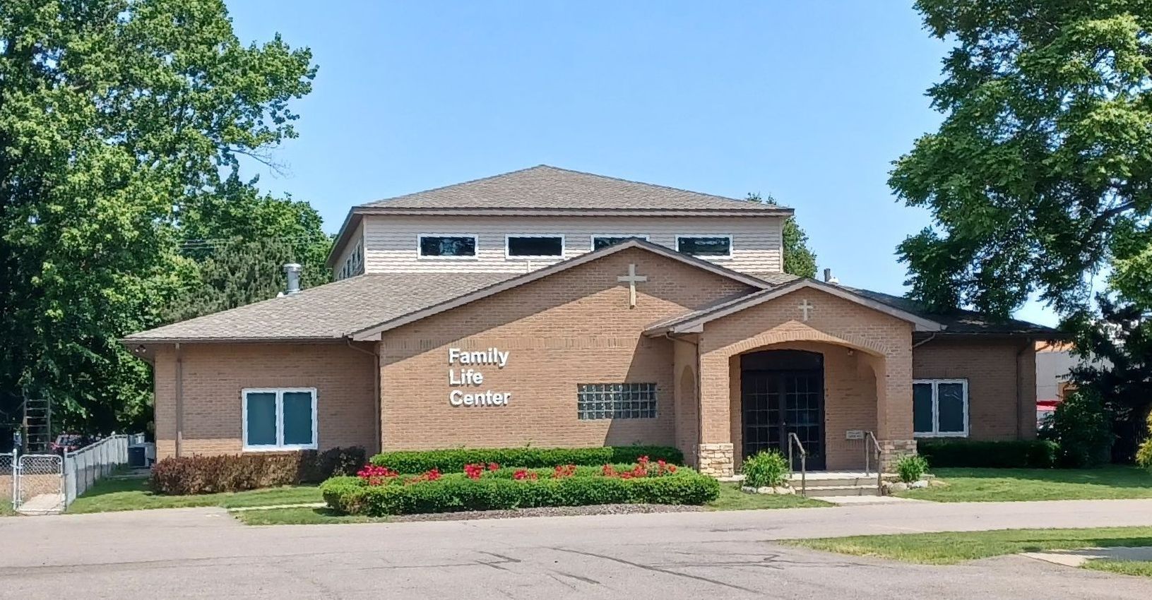 Tan brick church building with cross on roof, arched entrance, and a flower bed.