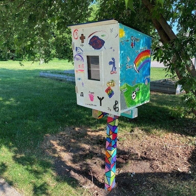 Colorful, decorated free library box on a painted post in a grassy area with a tree.