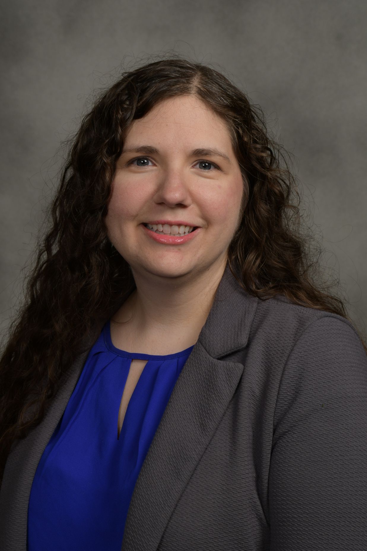 Woman with long curly brown hair wearing a gray blazer and blue top smiling against a neutral gray background.