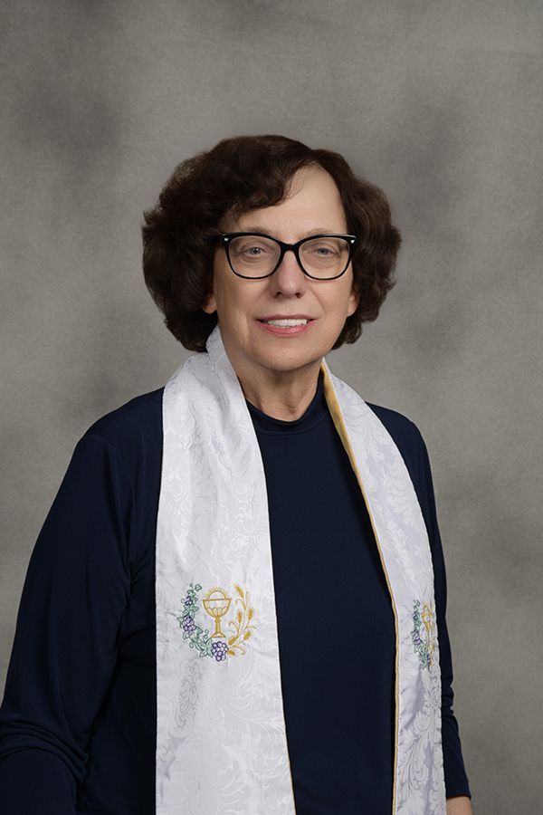 Woman in glasses, smiling, wearing a white stole with floral and chalice embroidery. Navy top, studio setting.