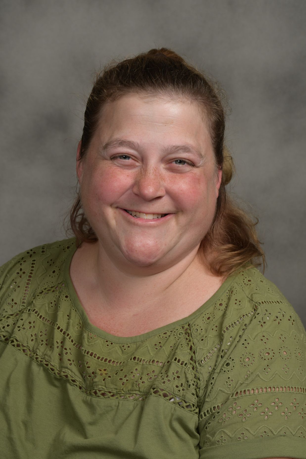 Woman with brown hair smiles, wearing a green eyelet top, against a gray background.