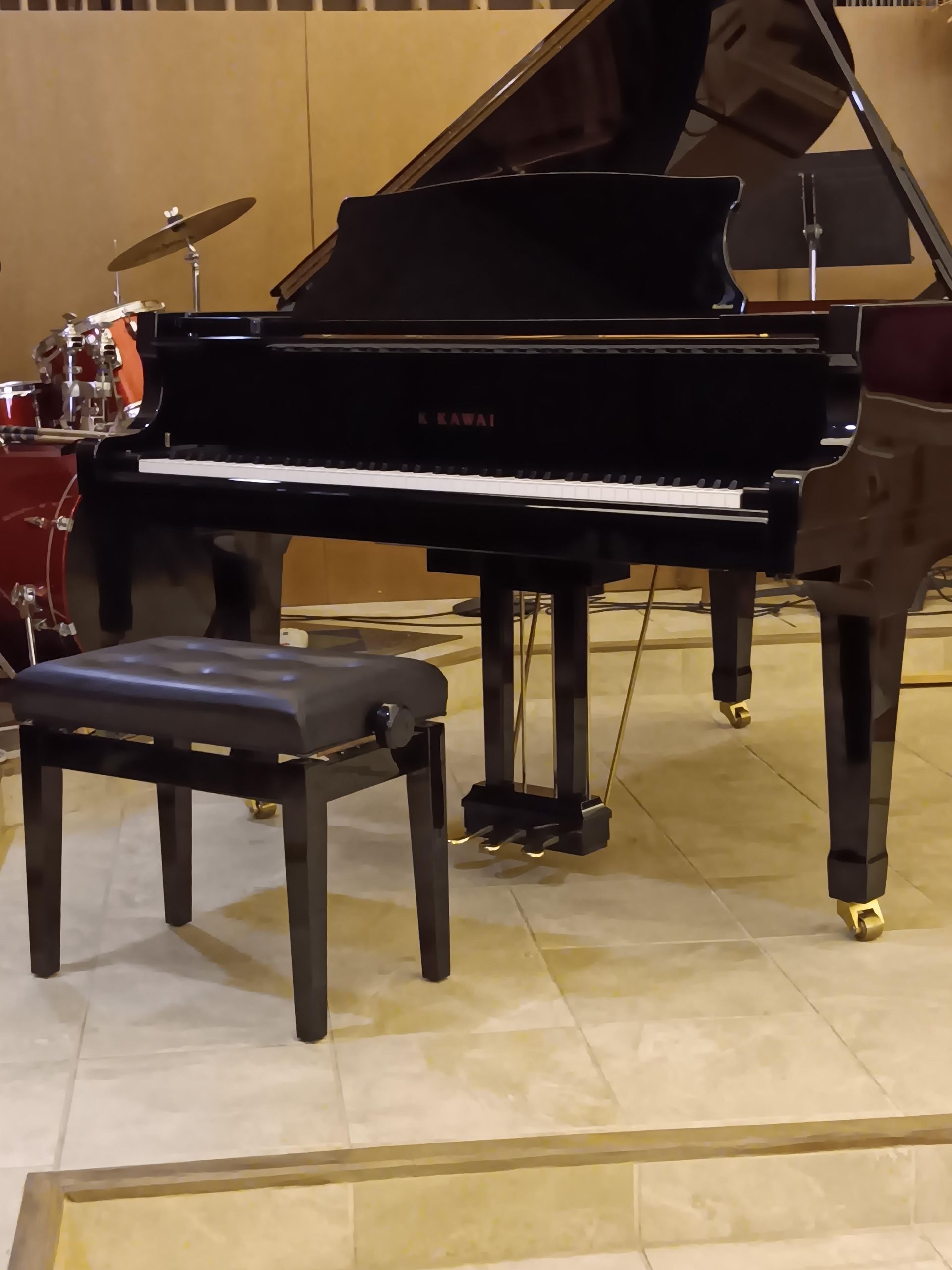 Black grand piano and adjustable stool in a room with a drum set visible in the background.