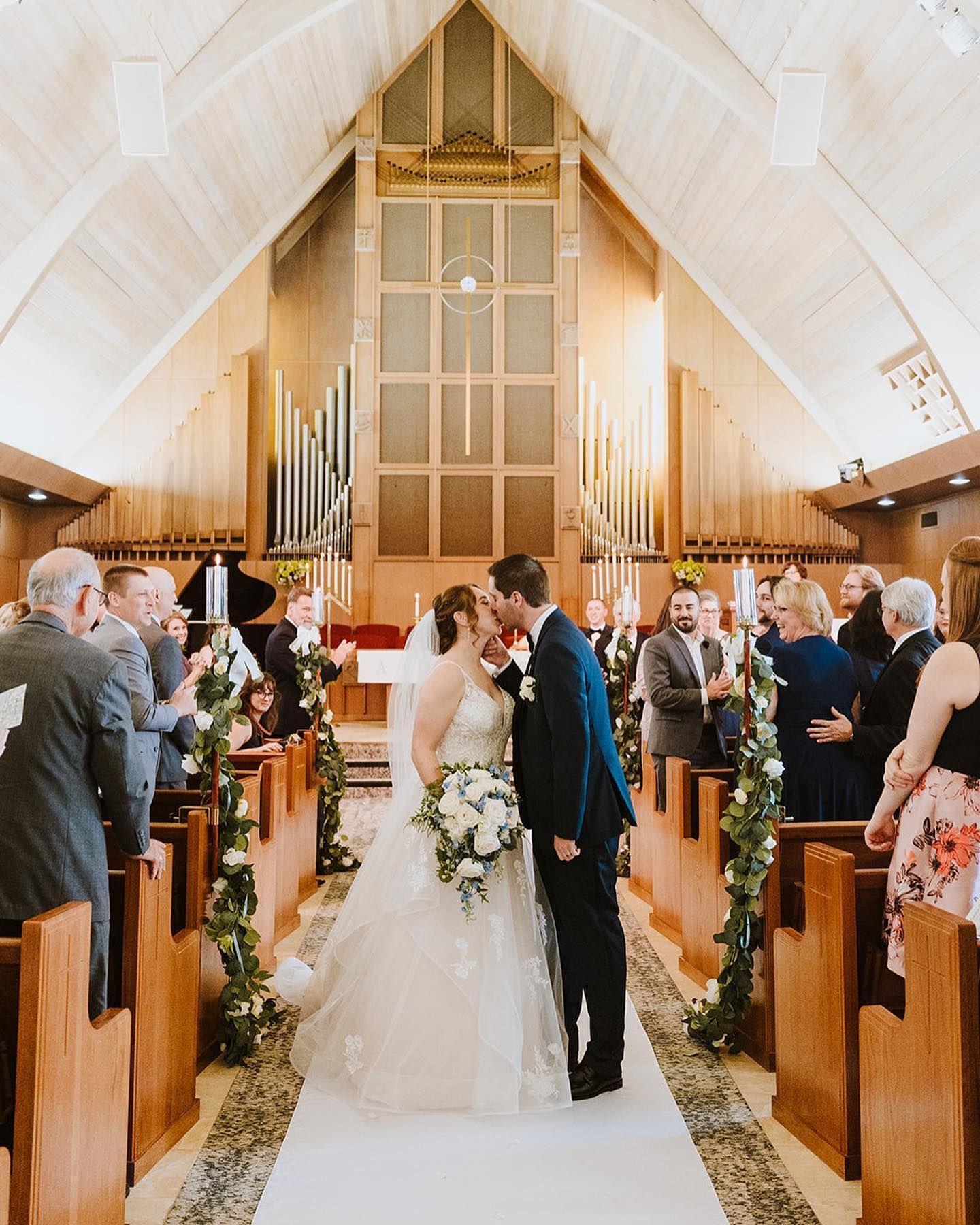 Bride and groom kiss at altar, wedding guests watch. Church with white aisle runner, floral garlands.