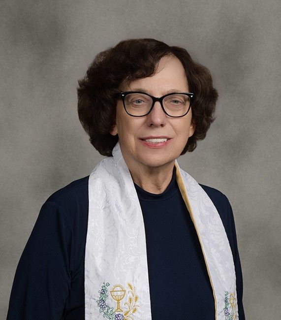 Woman wearing glasses and clergy stole, smiling, against a mottled gray background.