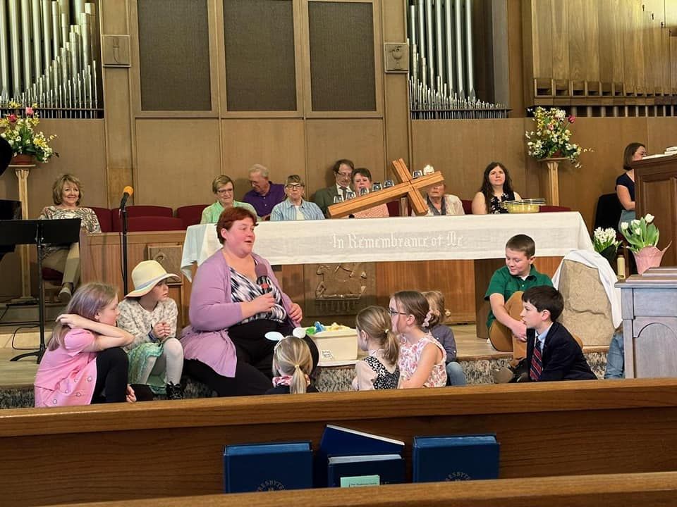 Church service with children. Woman speaks, other people sit/stand behind a table with a cross. Wooden pews in front.