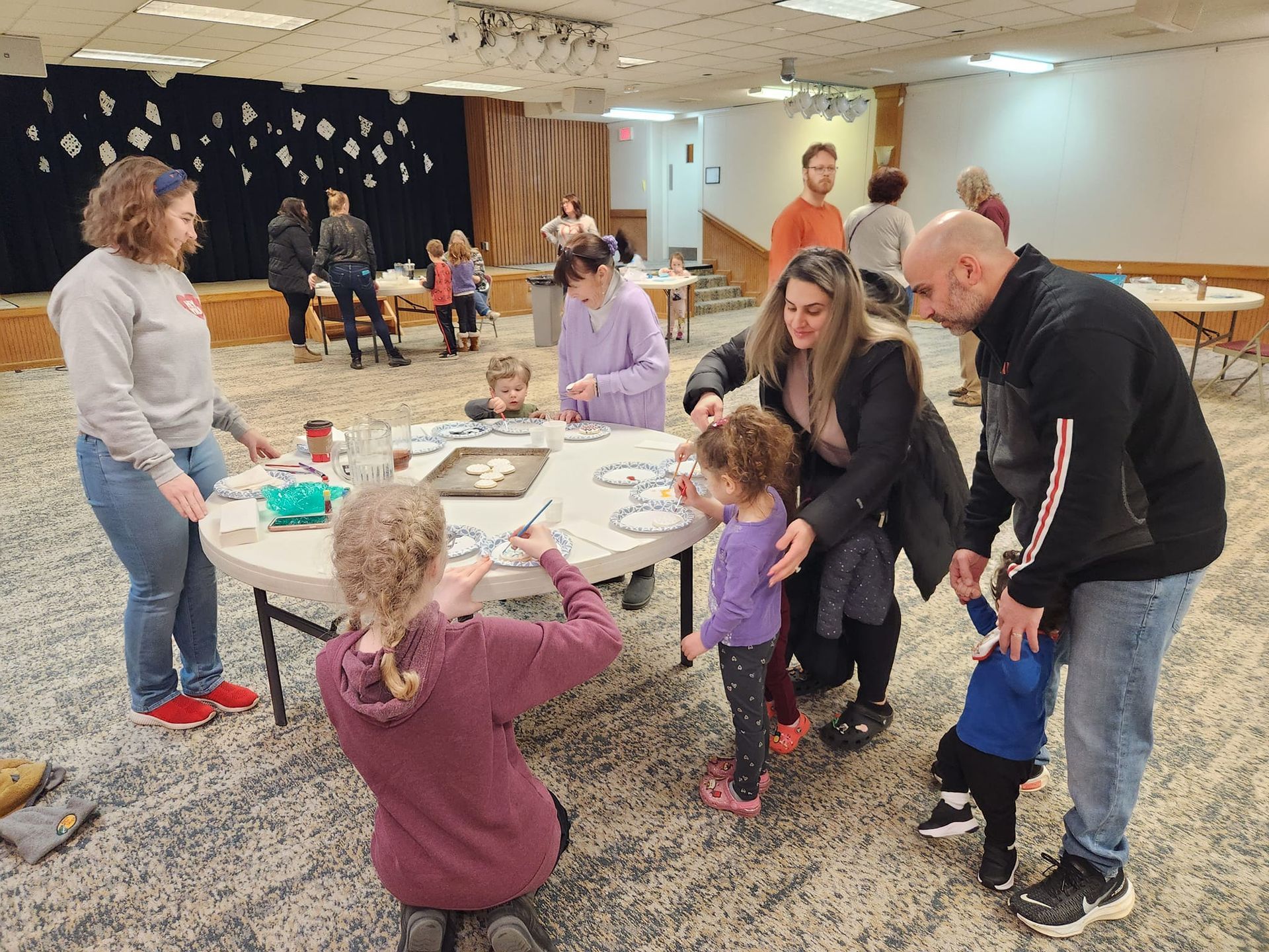 People decorate cookies at a table. Others watch, standing or sitting in a brightly lit room.