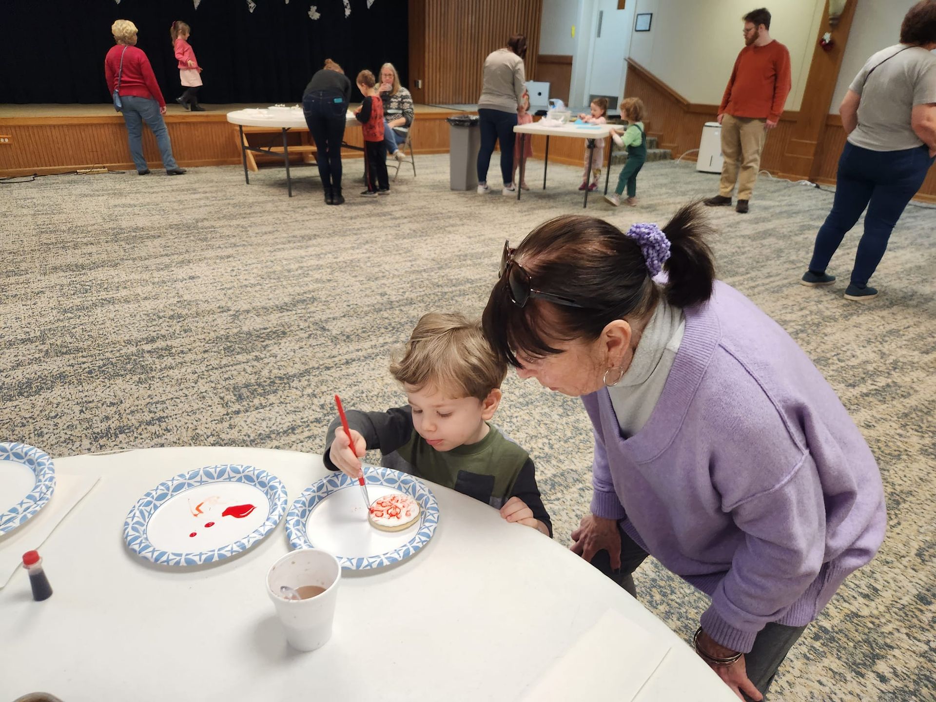 A child paints frosting on a cookie with an adult, at a craft table in a community hall.