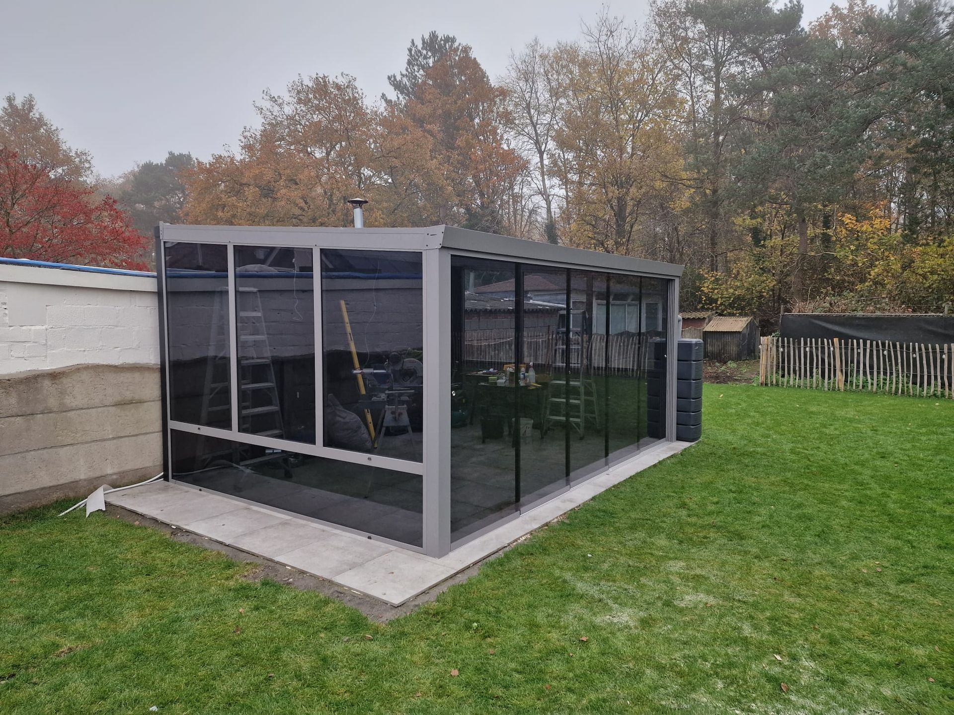 Moderne, zwart omlijste glazen serre, aangebouwd aan een huis, grenzend aan een grasveld en bomen in de achtertuin.