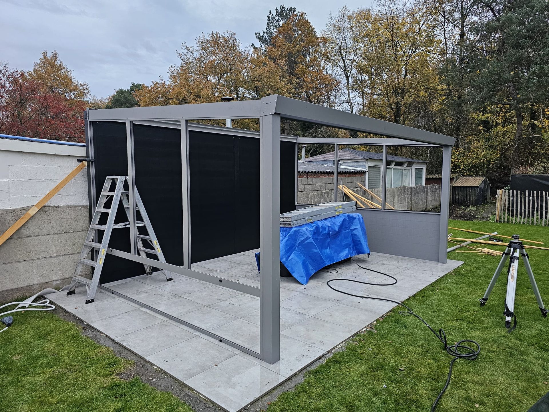 Een gedeeltelijk gebouwde grijze terrasconstructie met ladder, zwarte wandpanelen en een blauw zeil in een achtertuin.