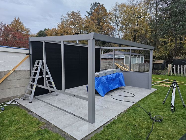 Een gedeeltelijk gebouwde grijze terrasconstructie met ladder, zwarte wandpanelen en een blauw zeil in een achtertuin.