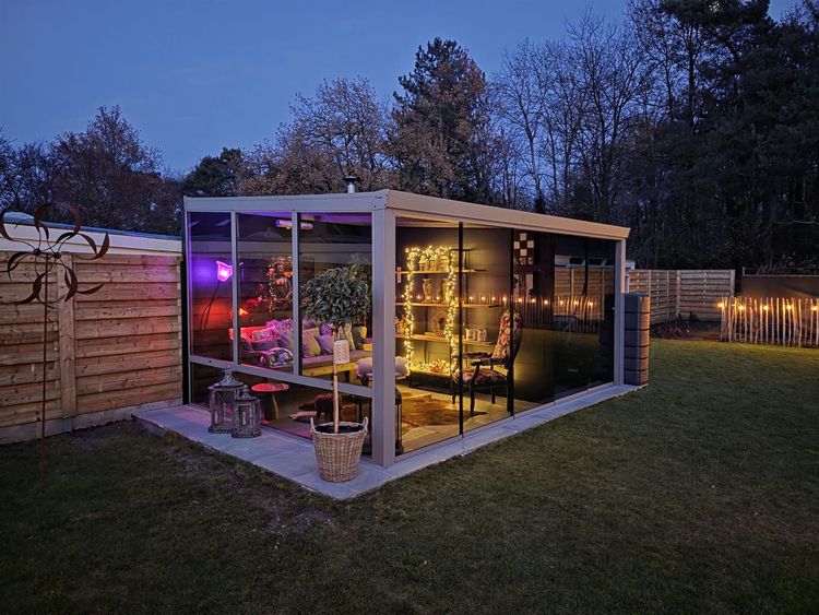 Een kleine, glazen tuinkamer in de achtertuin, die bij schemering baadt in warm licht, naast een houten schutting en gazon.