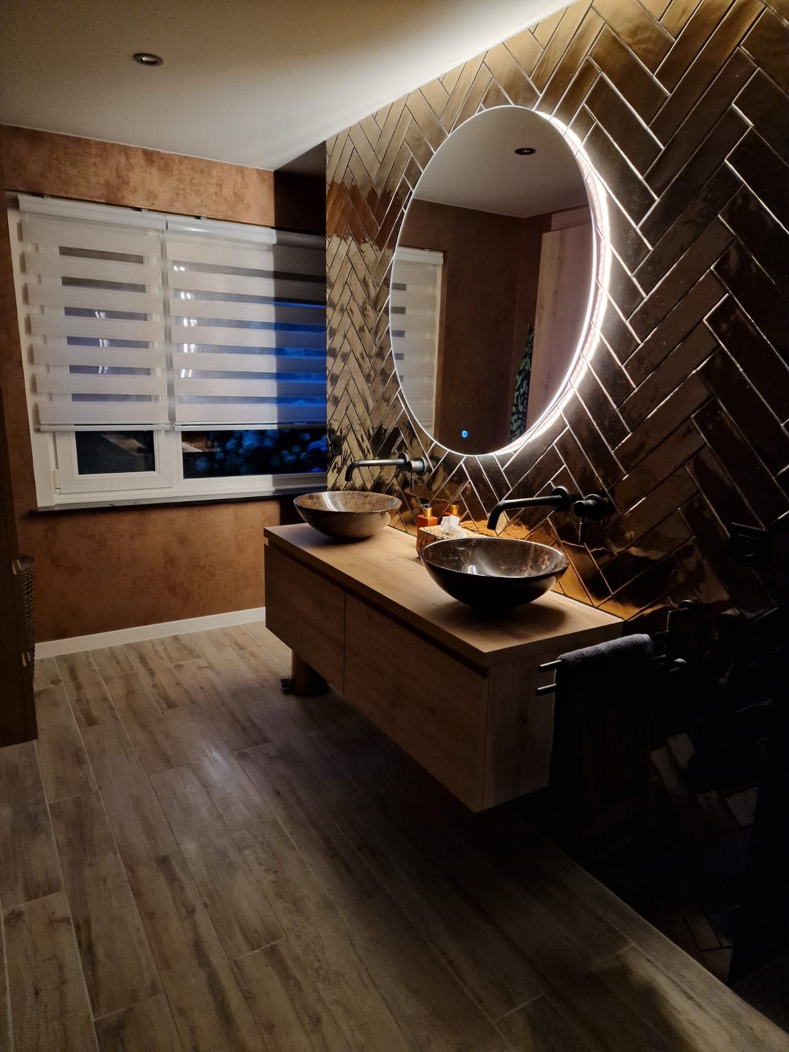 Moderne badkamer met een verlichte ronde spiegel, houten wastafelmeubel en raamjaloezieën.