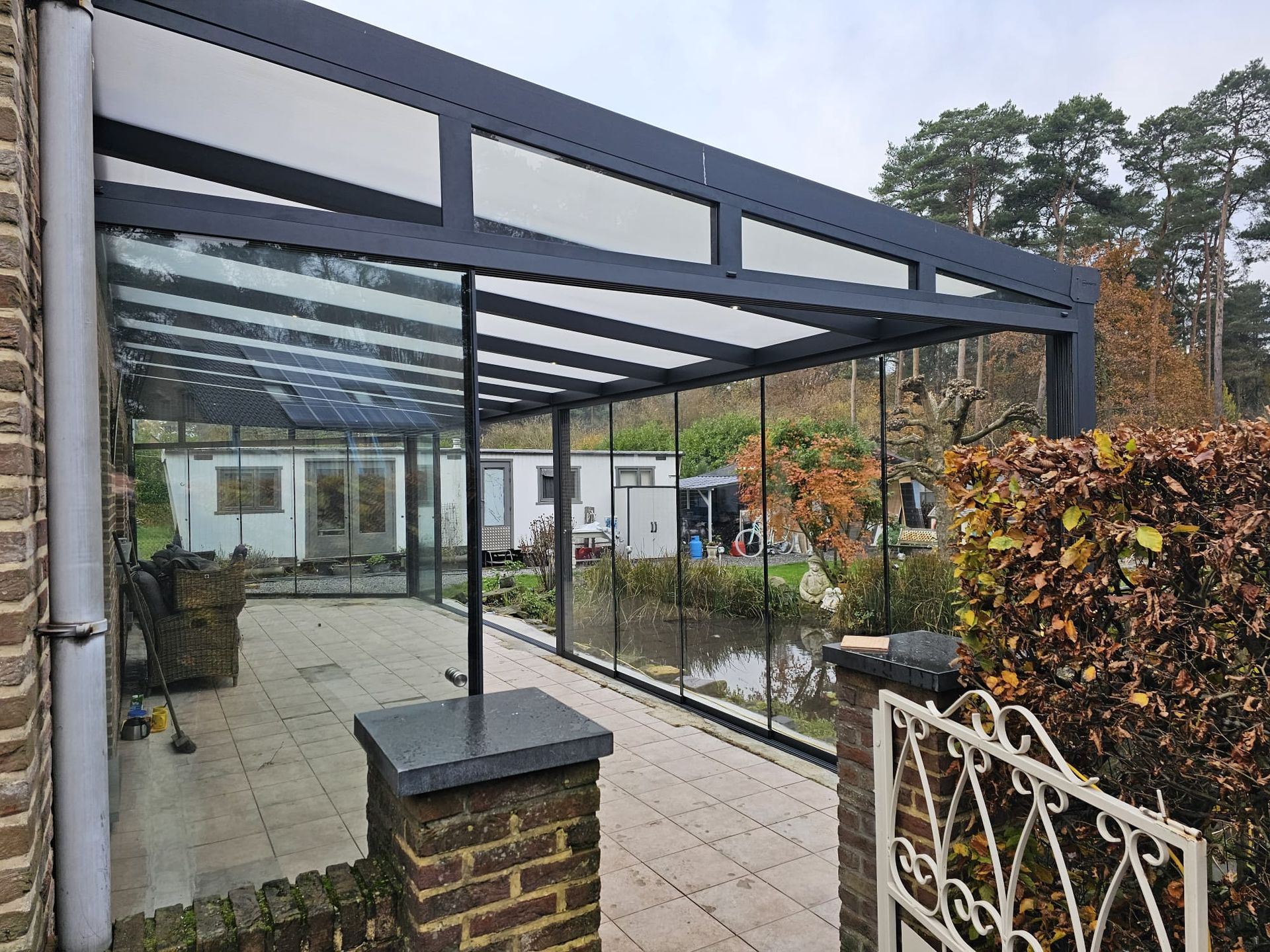 Overdekt terras met glazen dak en bakstenen pilaren naast een huis, dat uitkomt op een tuinpad.
