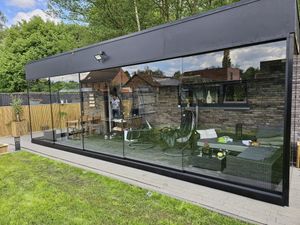 Moderne tuinkamer met glazen wanden en zwart frame, gelegen op een grasveld met weerspiegeling van bomen en huizen.