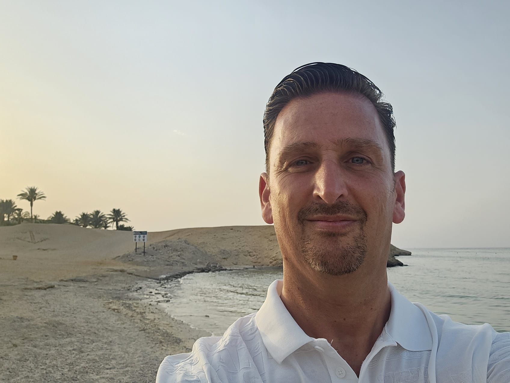 Man die een selfie maakt op een zandstrand bij zonsondergang, met palmbomen op de achtergrond.