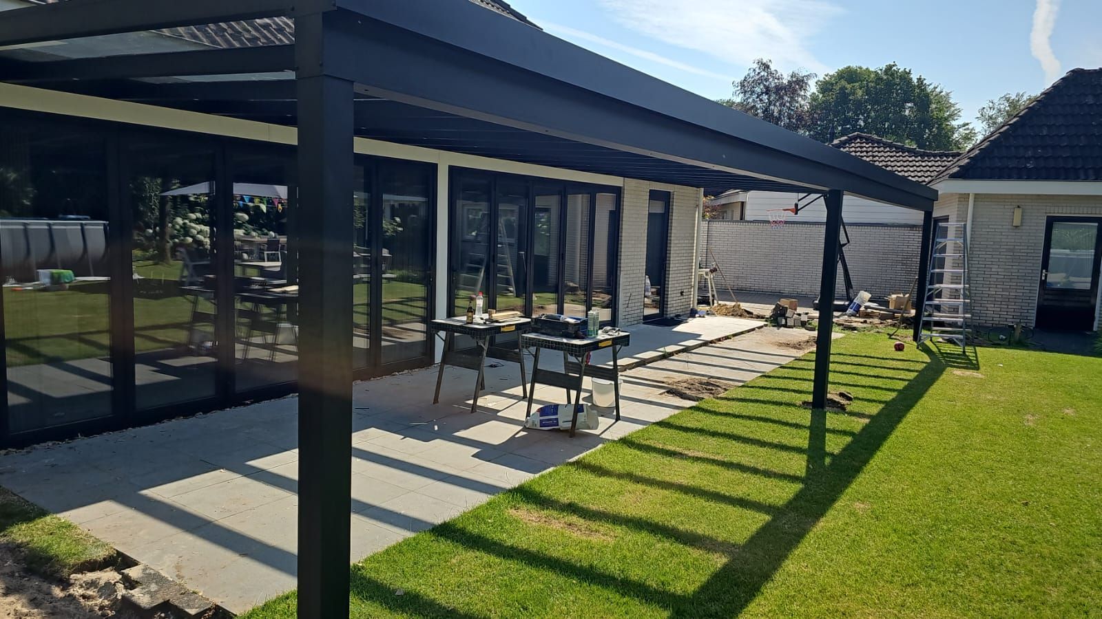 Terras met zwarte pergola, eettafel en loungestoelen op een zonnig groen gazon.