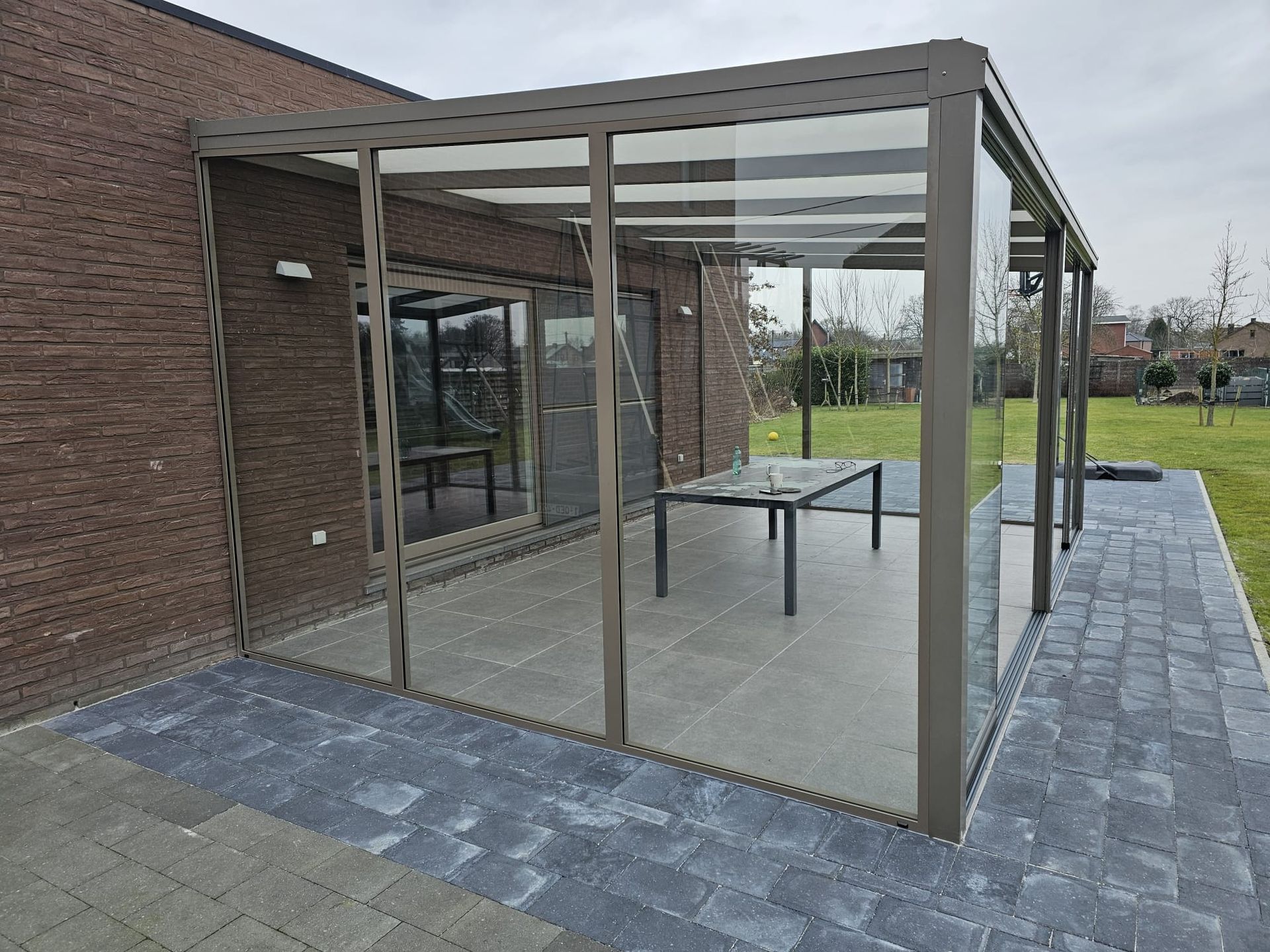 Glazen overkapping naast een bakstenen gebouw, met een tafel op een betegeld terras.
