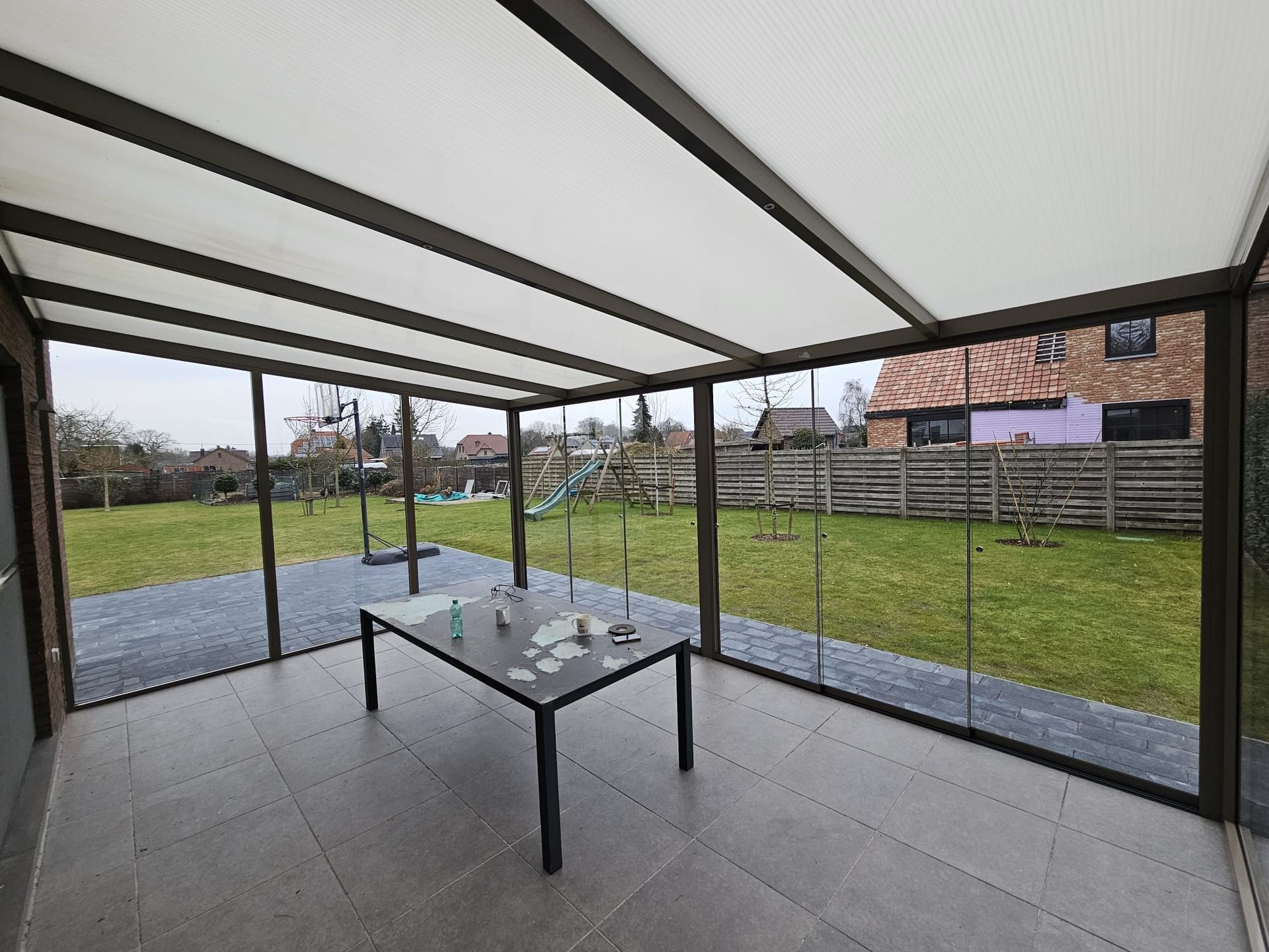 Overdekt terras met glazen wanden, tegelvloer en een tafel met uitzicht op een grasrijke achtertuin.