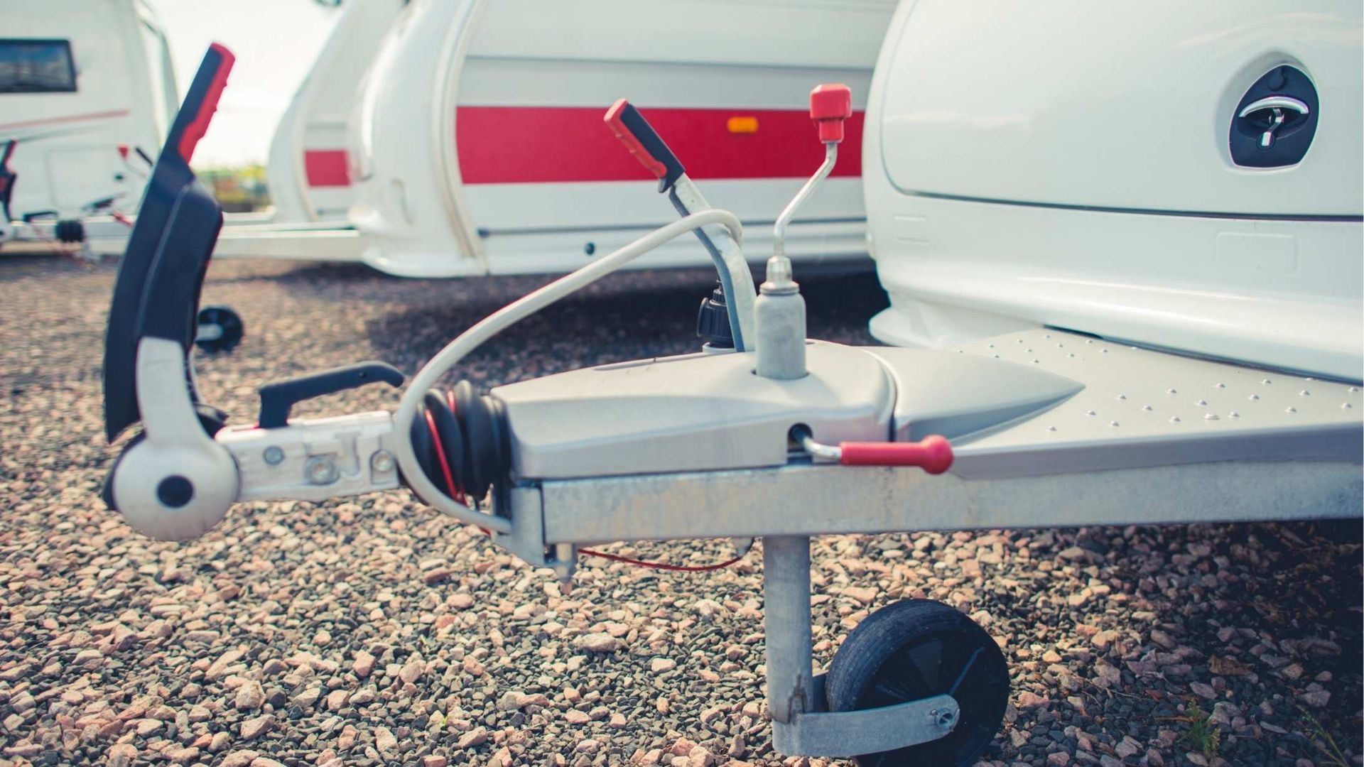 Close-up of a white caravan trailer hitch with a jockey wheel and red levers, parked on gravel with other trailers nearby.