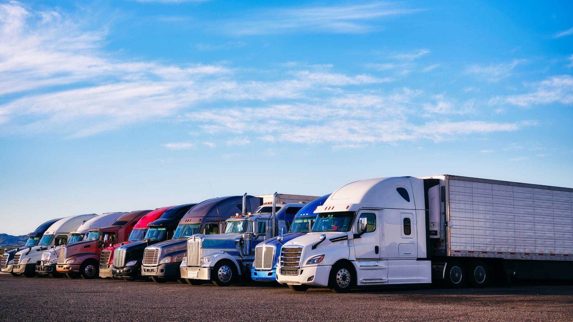 A row of semi-trucks with trailers parked on a gravel lot under a clear blue sky.