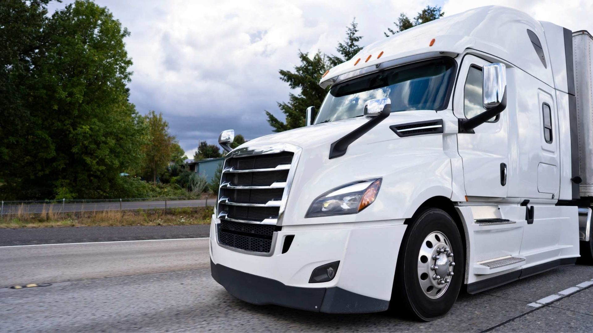 A white semi-truck driving on a road surrounded by trees under a cloudy sky.