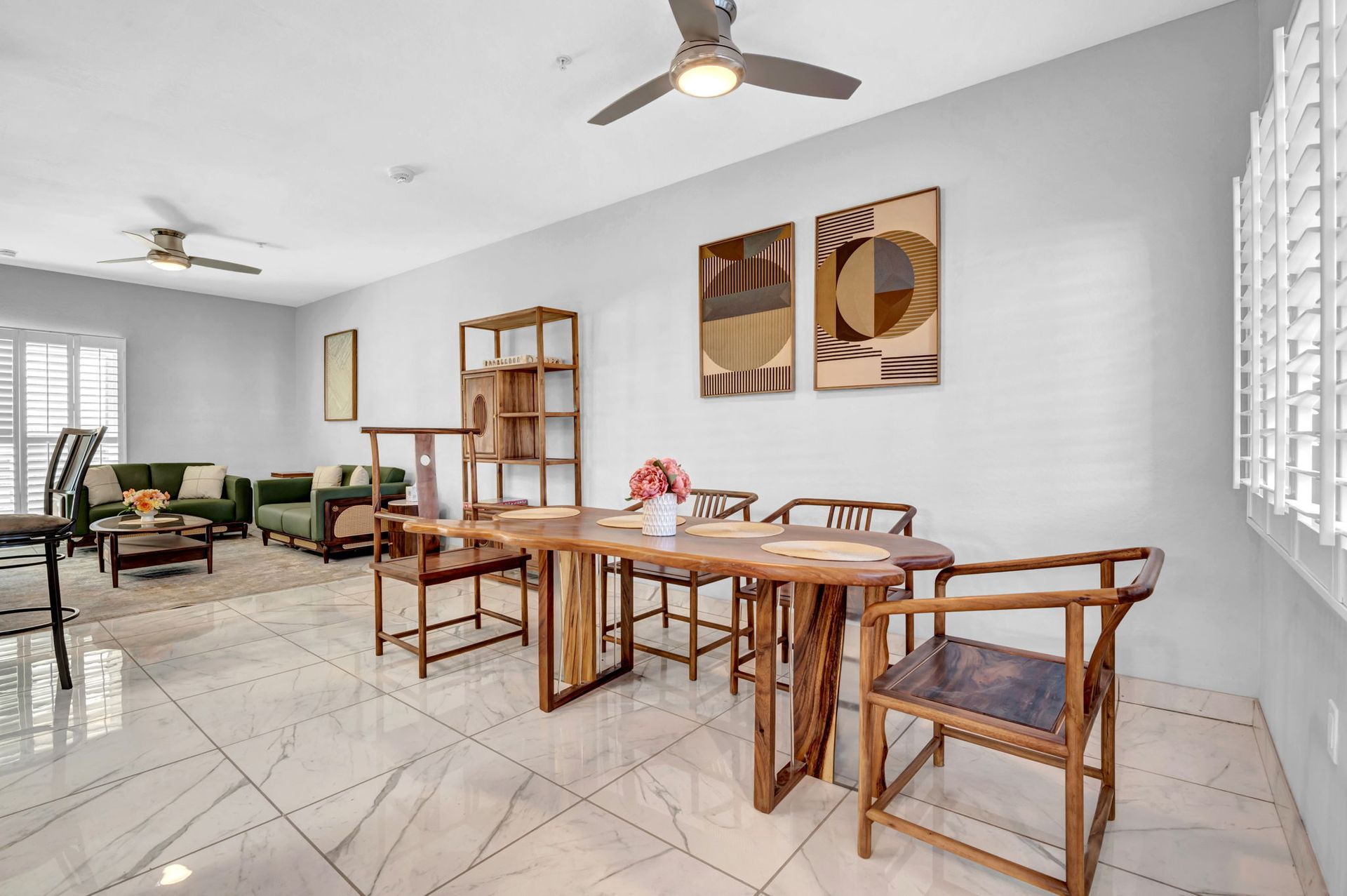Dining room with wooden table, chairs, and shelving. Artwork, ceiling fans, and green couch visible.