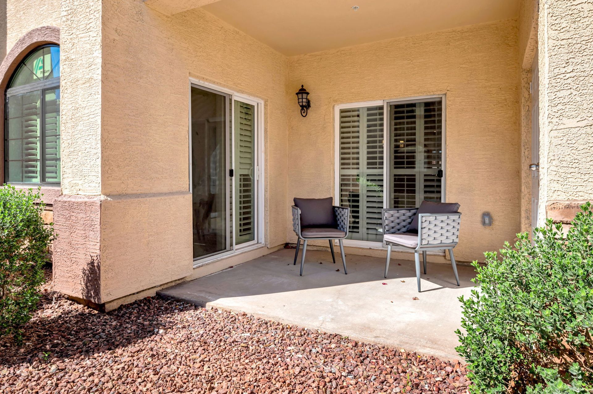 Patio with two chairs, sliding glass door, and a shuttered window. Beige walls and reddish-brown ground cover.