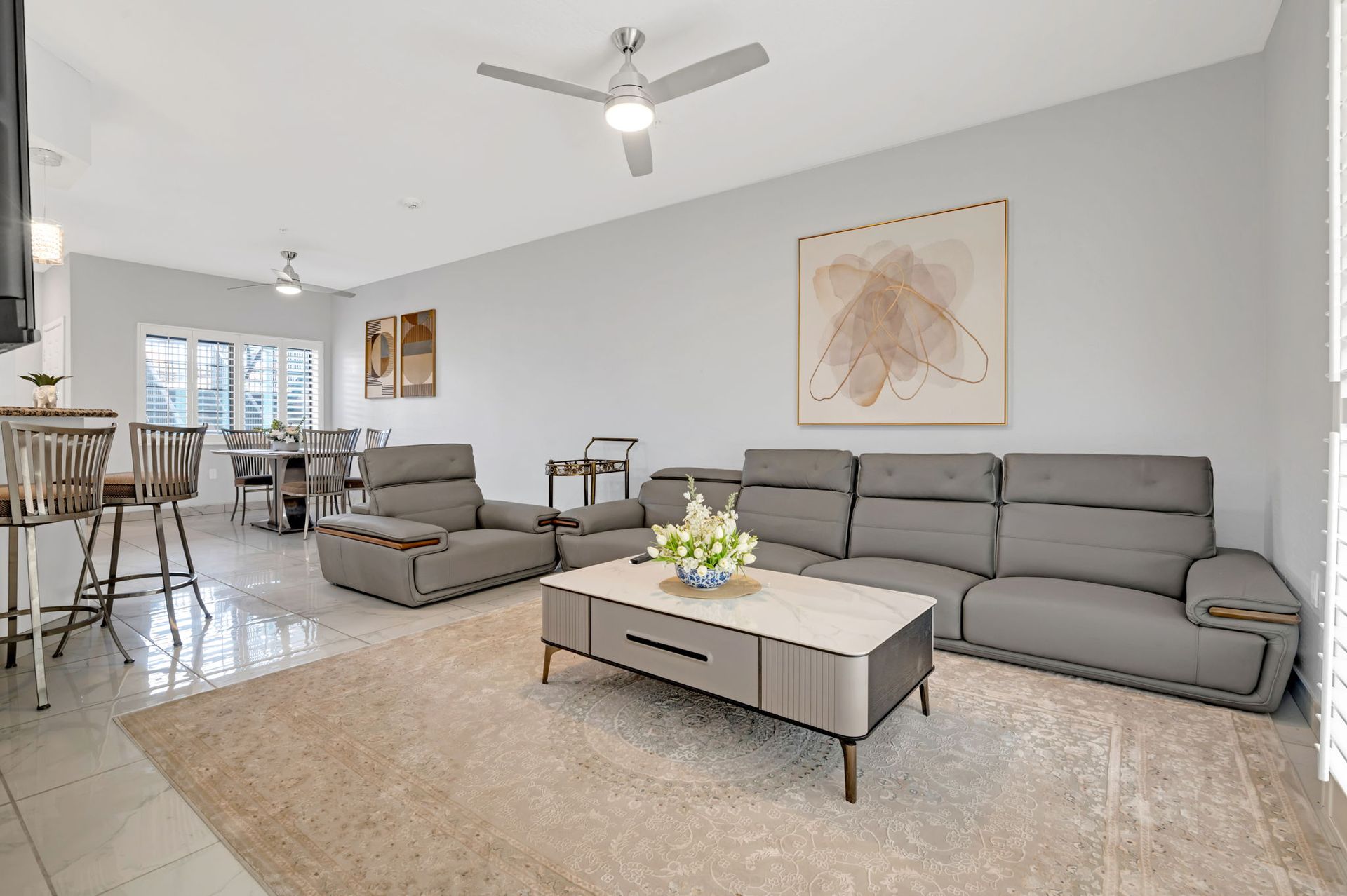 Modern living room with grey sofa, rug, and artwork. Dining area visible in the background.