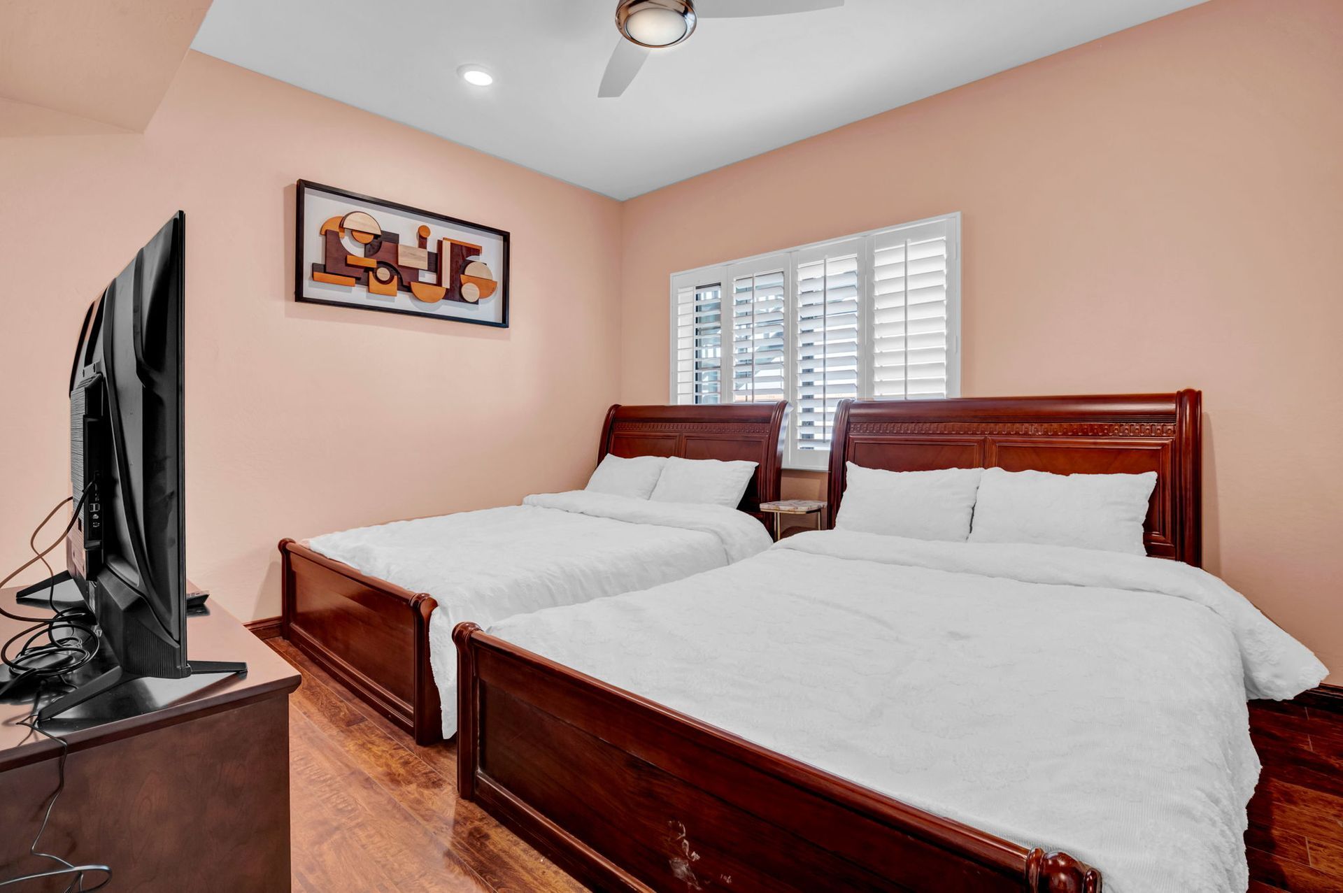 Bedroom with two beds, a TV, and a window with blinds. Pink walls, dark wood furniture.