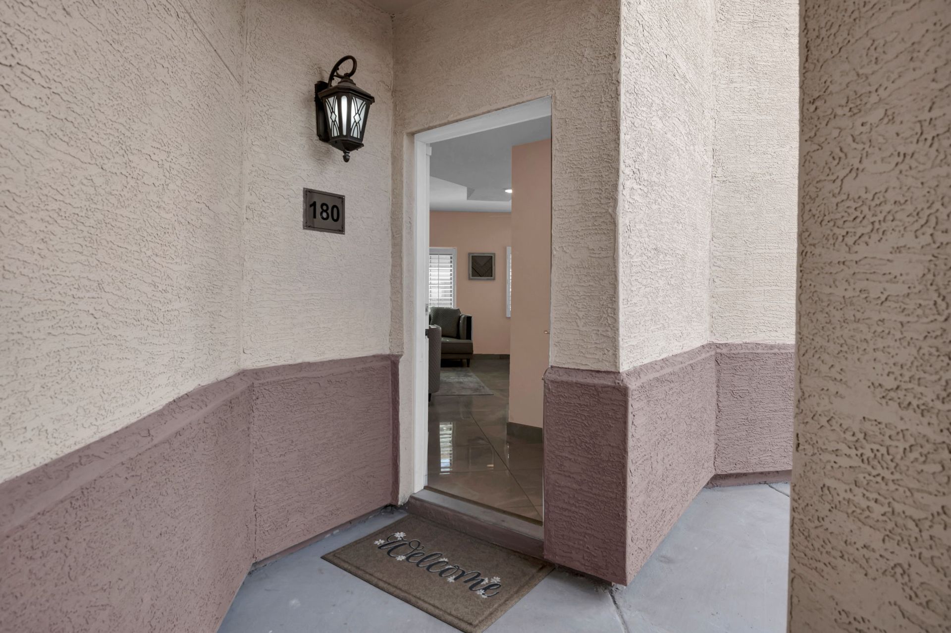 Exterior view of apartment doorway, light brown textured walls with a door mat reading 