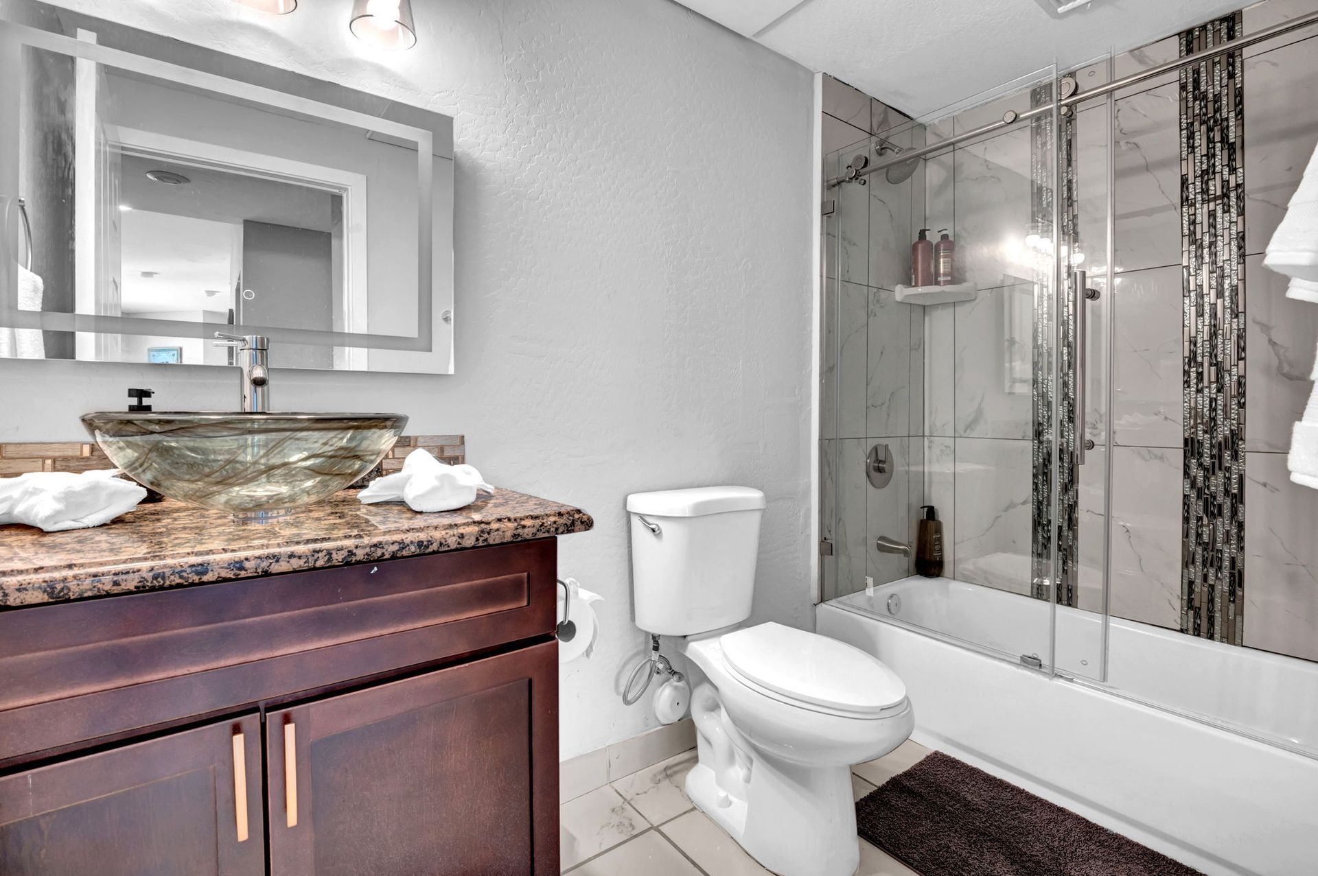 Bathroom with a dark wooden vanity, glass vessel sink, white toilet, and glass shower enclosure.