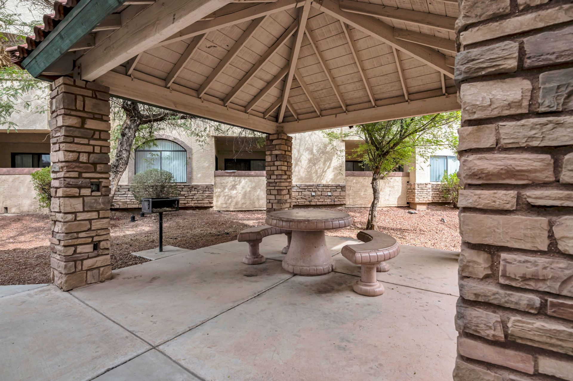 Covered patio with stone columns, concrete table, and benches; a grill in the background.