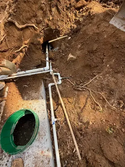 Exposed PVC pipes in a dirt excavation beside a green drain opening and concrete slab