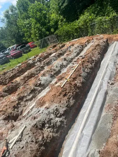 Prepared soil rows covered with plastic sheeting on a sloped hillside, with parked cars and trees nearby
