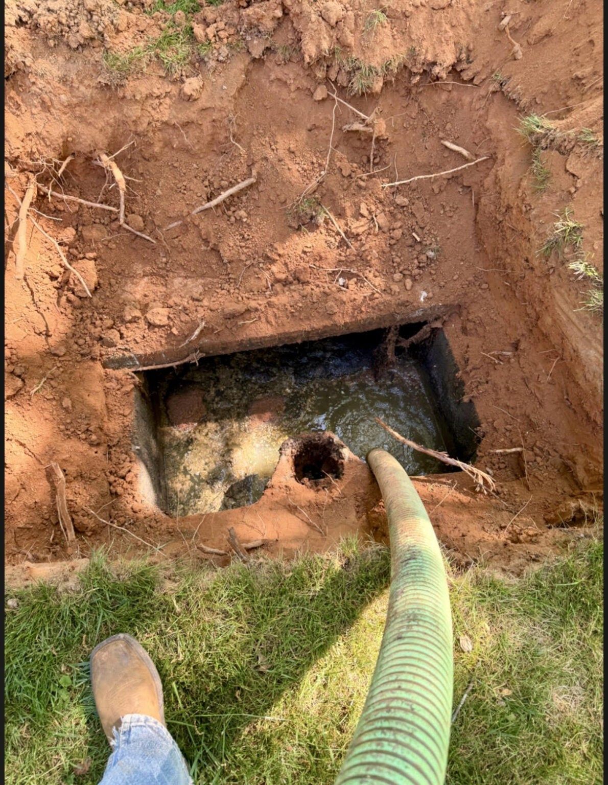 A dirt hole with a green corrugated hose inserted into a buried opening, next to a grassy edge.