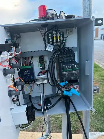 Open electrical control panel with wiring, relays, and components mounted outdoors on a gray box.