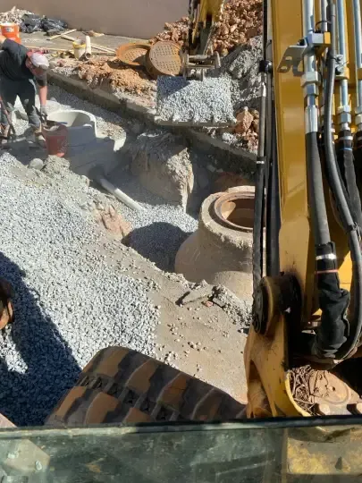Construction site with excavator arm and worker shaping a gravel trench around concrete pipes.