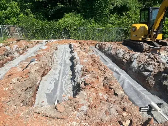 Excavated trenches lined with plastic sheeting at a construction site, with an excavator on the right.