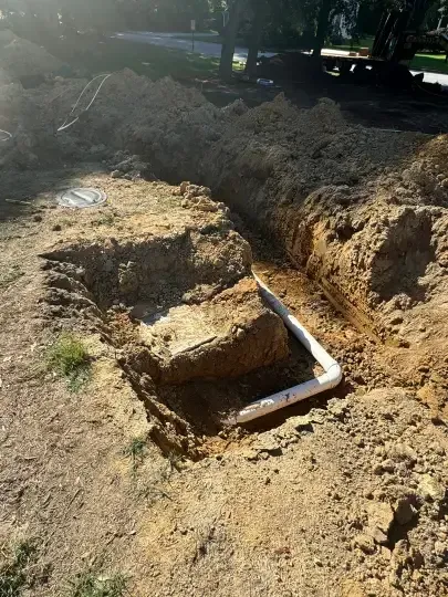 Excavation trench with exposed concrete footing and white pipe in a dirt lot near trees