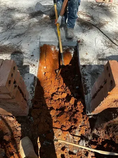 Worker digging a narrow trench beside stacked bricks, with a shovel and muddy soil.